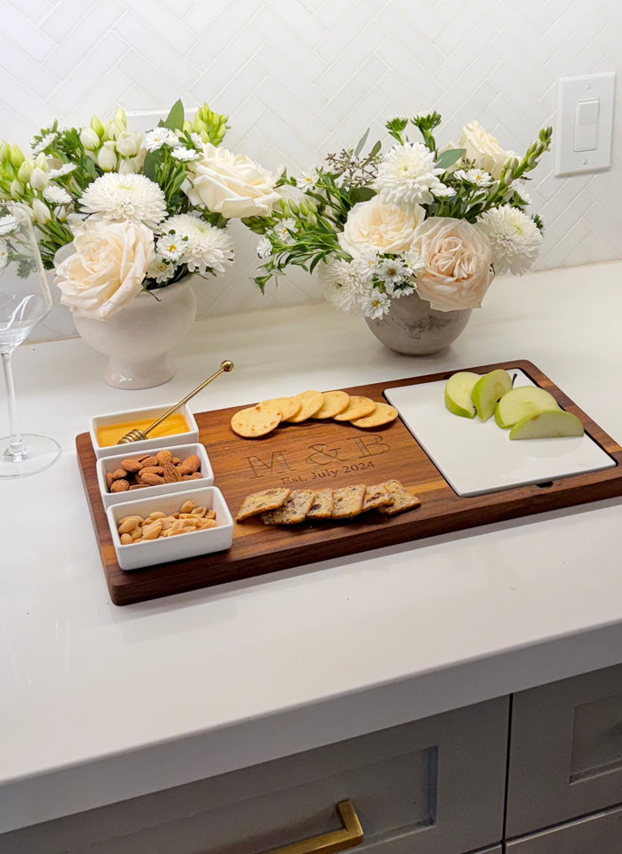 The Maple Acacia Fête Set Tray (18 x 9.5, personalized) is displayed on a white countertop with crackers, apple slices, mixed nuts, and snacks. Ceramic snack dishes, vases with white flowers, and a wine glass are in the background.