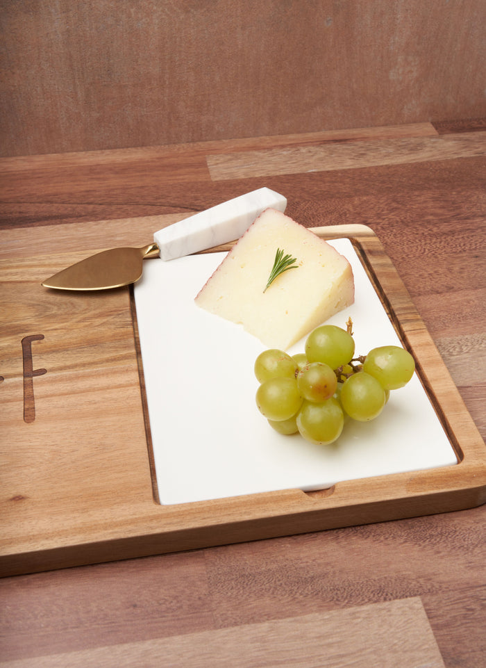 A wedge of cheese with rosemary and green grapes sits on the Maple Acacia Fête Set Tray (Personalized, 18 x 9.5) with a gold cheese knife, all arranged on a wooden surface.