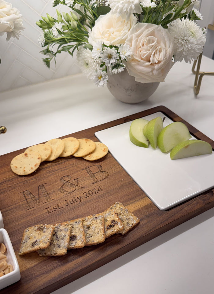 The Maple Acacia Fête Set Tray (18 x 9.5), personalized with "M & B Est. July 2024," displays crackers, green apple slices, and nut bars. Ceramic snack dishes and a vase of white flowers add elegance in the background.