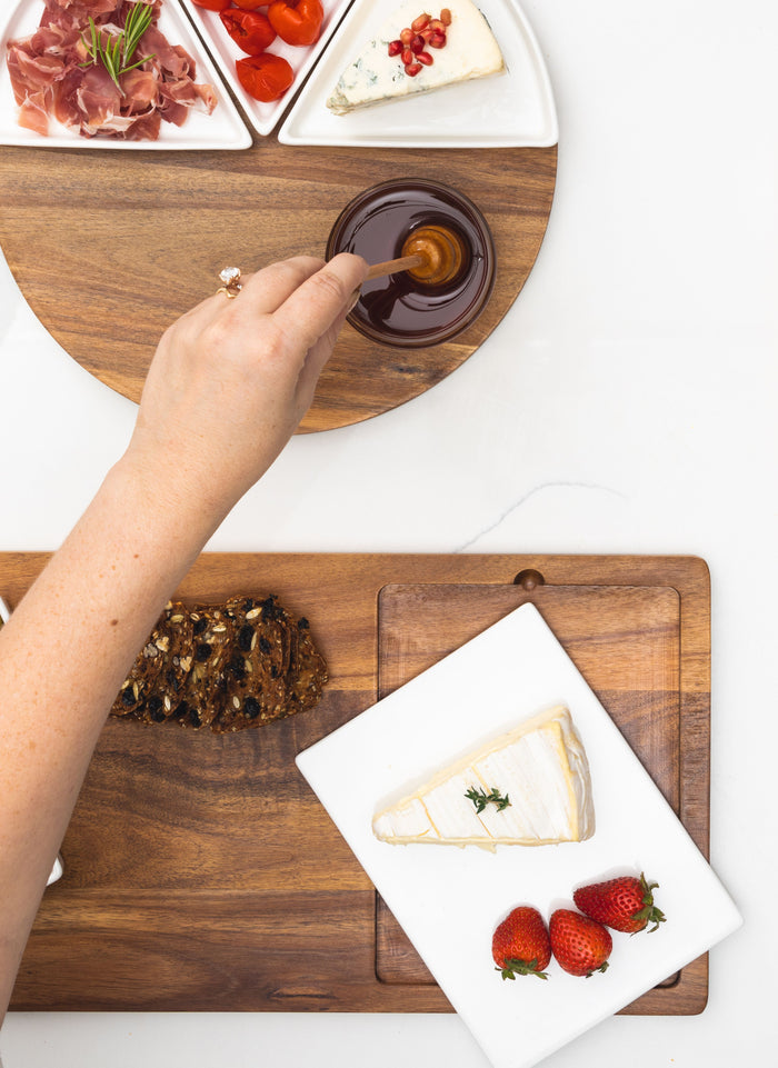 A hand dips a honey dipper into honey on the Maple Acacia Fête Set Tray (Personalized, 18 x 9.5), surrounded by cheese, cured meat, cherry tomatoes, crackers, and strawberries.
