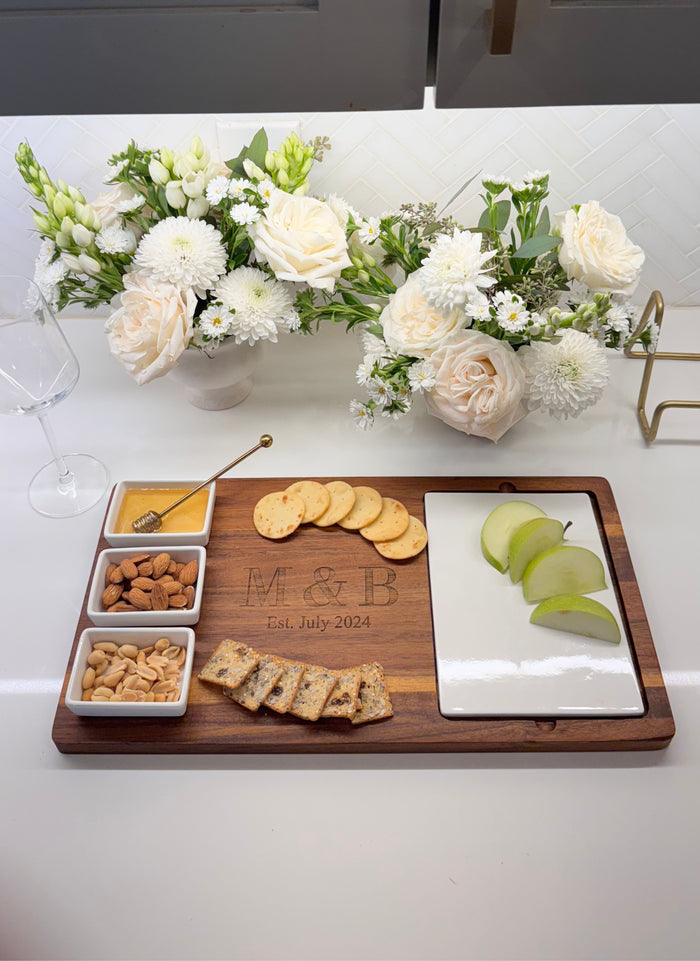 The Maple Acacia Fête Set Tray (Personalized, 18 x 9.5) is topped with crackers, green apple slices, nuts, and honey on a counter. Behind it are two floral arrangements of white and pale pink blooms and an empty wine glass.