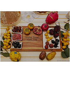An Acacia Fête Set Tray by Maple (18" x 9.5") rests on a striped table, showcasing assorted exotic fruits—dragon fruit, persimmon, pomegranate, yellow plums, figs, and dates—with a cake in the corner.