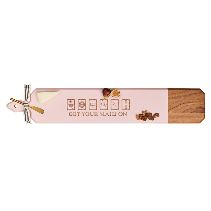 The Maple Acacia Heirloom Board Skinny in pink, featuring a wood design and engraved with "Get Your Mahj On" mahjong tiles (34 x 6"), includes a gold spreader and ribbon—perfect for serving cheese, figs, and nuts at your next gathering.