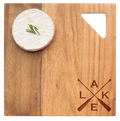 A round wheel of brie with chives sits on the Maple Acacia Modern Geo Square | Lake | 9 x 9" board, which features a triangle cutout and crossed paddle engraving with L, A, K, and E in the lower right corner.
