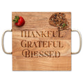 The Maple Acacia Modern Square Charcuterie Board (12" x 12") features gold handles and "Thankful Grateful Blessed" engraving, shown with a strawberry and two pieces of fruit and nut crispbread on top.