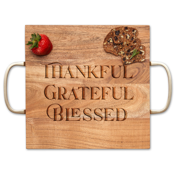 The Maple Acacia Modern Square Charcuterie Board (12" x 12") features gold handles and "Thankful Grateful Blessed" engraving, shown with a strawberry and two pieces of fruit and nut crispbread on top.