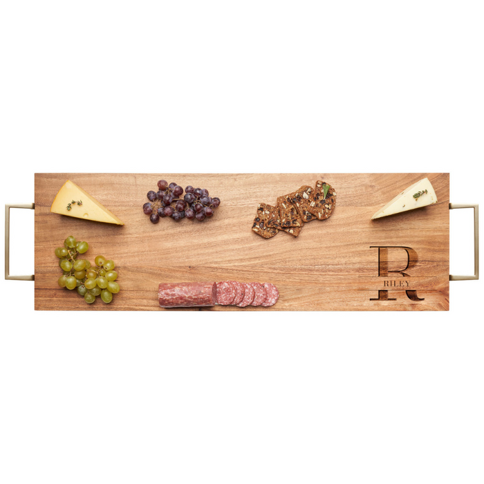 The Maple Acacia Serving Board with Gold Handles (30 x 10") displays two cheese wedges, green and red grapes, crisp crackers, and sliced salami. This elegant board is engraved with a large letter R and the name Riley.