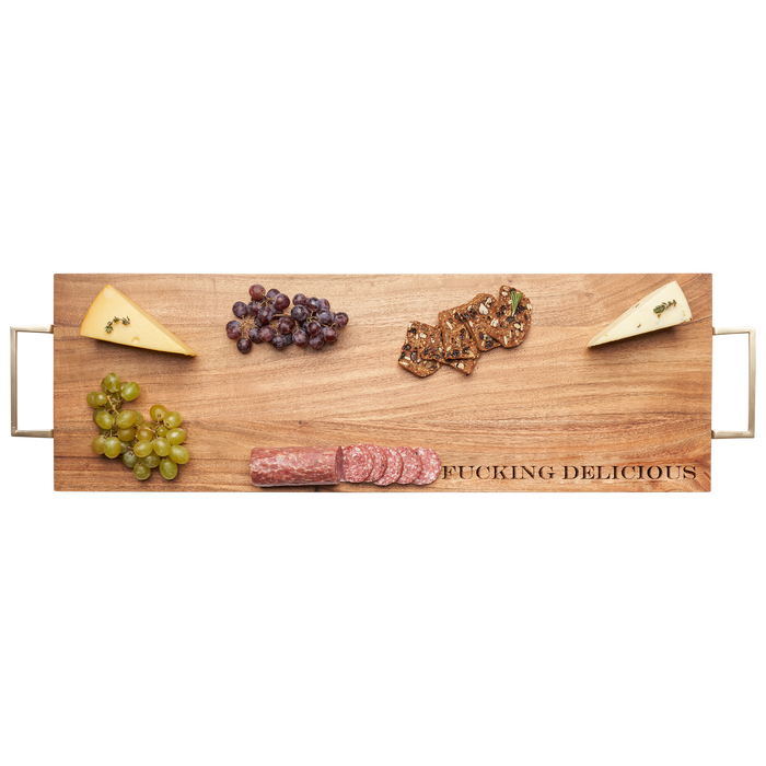 The Maple Acacia Serving Board (30 x 10") with gold handles features "Fucking Delicious" engraved and comes arranged with two cheeses, green and red grapes, crackers, and sliced salami.