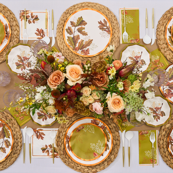 A fall-themed table setting features floral centerpieces, woven placemats, leaf-printed plates from the Acorn and Oak Collection, and gold-accented cutlery on a white tablecloth for an elegant and festive atmosphere. The arrangement is completed with Sophistiplate’s Acorn and Oak Appetizer/Dessert Bowls (8pk), which add both convenience and charm to this autumn-inspired display.