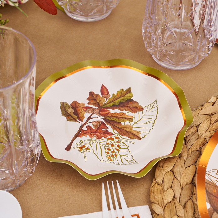 A Sophistiplate Acorn and Oak Collection scalloped paper plate, featuring a gold rim and adorned with autumn leaves, acorns, and flowers, rests on a brown table beside clear glasses, a woven placemat, and white plastic cutlery—perfect for Thanksgiving gatherings or pairing with Acorn and Oak Appetizer/Dessert Bowls (8pk).