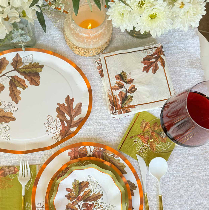 Table set with autumn-themed plates and napkins featuring brown leaves from the Acorn and Oak Collection, white utensils, a glass of red wine, a lit candle, and white flowers in a vase on a white tablecloth. For an inviting seasonal touch, add Sophistiplate’s Acorn and Oak Appetizer/Dessert Bowls (8 pack), perfect for serving fall treats at your Thanksgiving gathering.