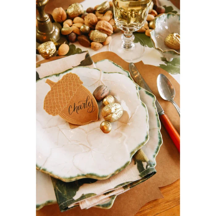 A festive Thanksgiving table features Hester & Cook’s Acorn Place Card - Pack of 12, layered leaf plates, a napkin, assorted nuts, gold-wrapped chocolates, cutlery, and a wine glass atop an autumn-themed tablecloth—perfect for celebrations.