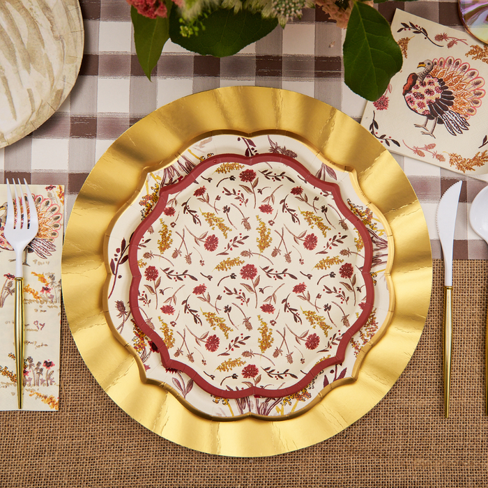 A decorative place setting featuring a gold charger, floral-patterned plate, white utensils with gold handles, napkins, and a bird-adorned paper accent—all arranged on a plaid and burlap tablecloth—creates an inviting tablescape. For added elegance and convenience, incorporate the Sophistiplate Amber Harvest Salad Plates (8-pack) as stylish disposable Thanksgiving plates to complete your festive look without compromising on style.