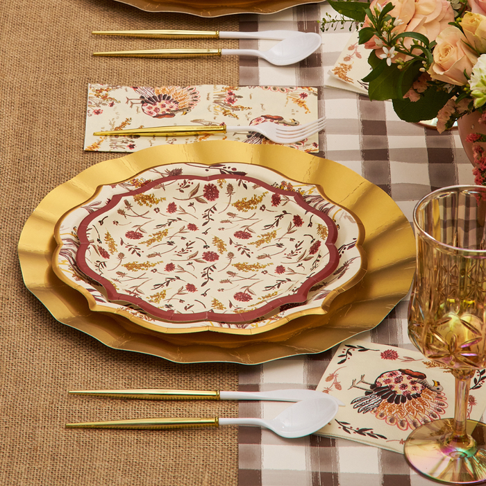 A table setting with Sophistiplate Amber Harvest Salad Plates (8pk), gold utensils, fancy-edge paper plates, plastic spoons, a plaid tablecloth, napkins with floral designs, and a bouquet of flowers in the corner.
