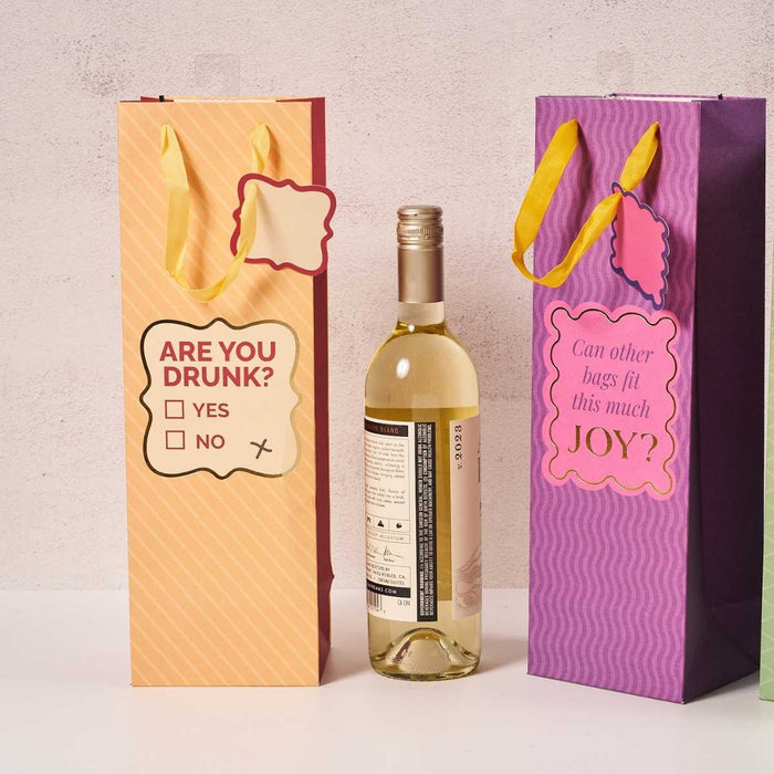A wine bottle sits between two vibrant gift bags from Drinks On Me. The Are You Drunk- Wine Bag is yellow with "ARE YOU DRUNK? YES/NO," while the purple bag reads "Can other bags fit this much JOY?" Both feature yellow handles and are reusable.
