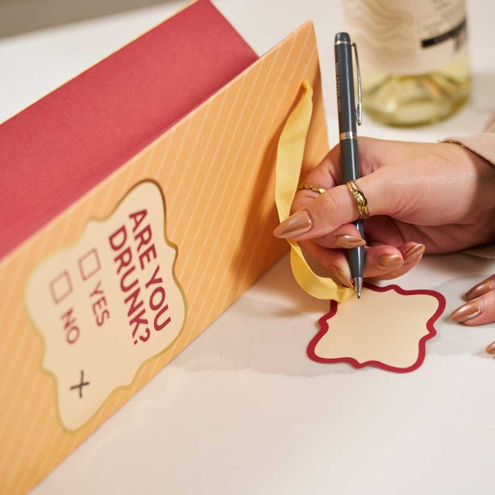 A manicured hand writes on a tag beside the Drinks On Me "Are You Drunk?" wine bag, featuring YES/NO checkboxes—a fun hostess gift. A wine bottle and glass blur in the background.