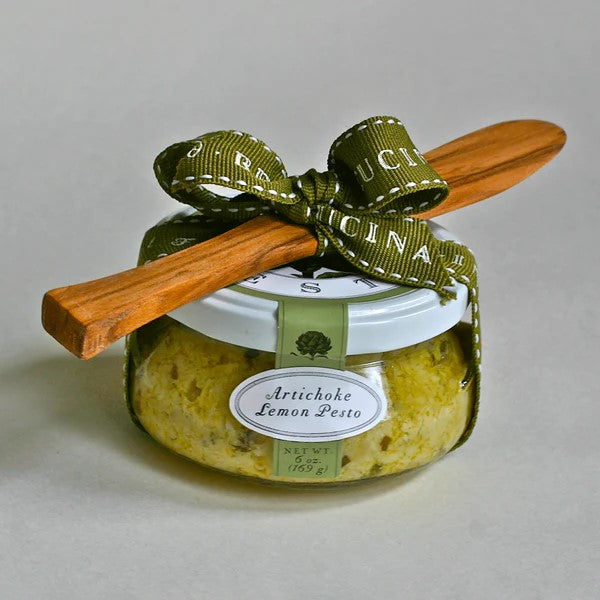 A glass jar of Bella Cucina Artichoke Lemon Pesto Gift Set features a white lid, green ribbon bow, wooden spreader, and a label displaying the product name—an Oprah’s Favorite Things pick.