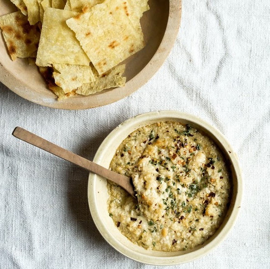The Bella Cucina Artichoke Lemon Pesto Gift Set, one of Oprah's Favorite Things, features a creamy dip garnished with herbs and served with a wooden spoon, paired perfectly with flatbread on a light textured cloth.