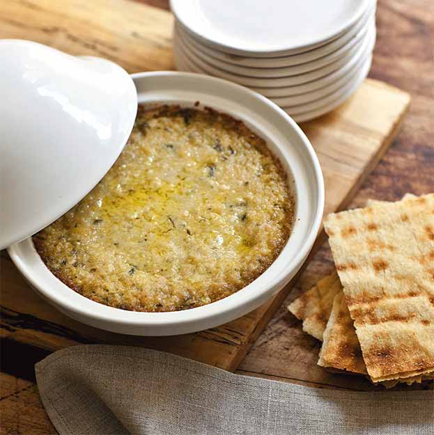 A round white dish of warm, baked Artichoke Lemon Pesto from Bella Cucina sits partially covered on a wooden board beside stacked white plates and flatbread, with a beige napkin draped in front.
