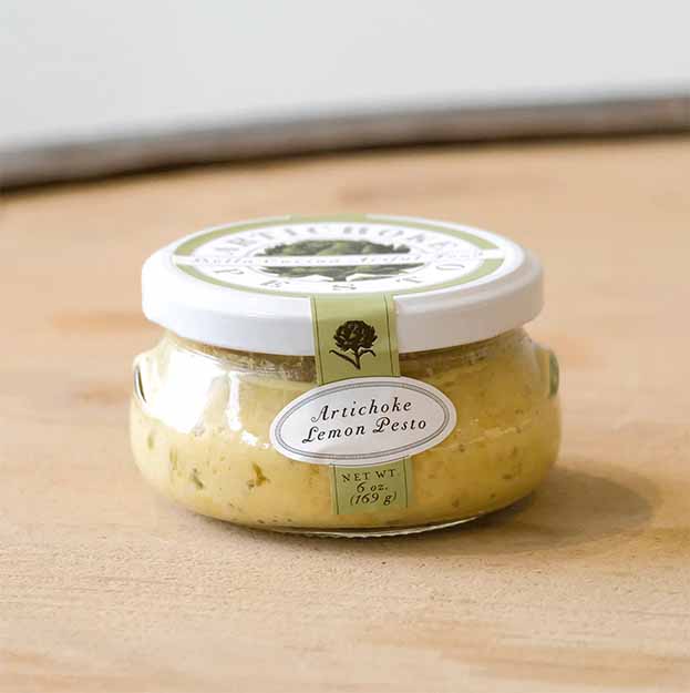 A small glass jar with a white lid labeled "Artichoke Lemon Pesto" by Bella Cucina holds a creamy yellow-green spread and sits on a light wooden surface.