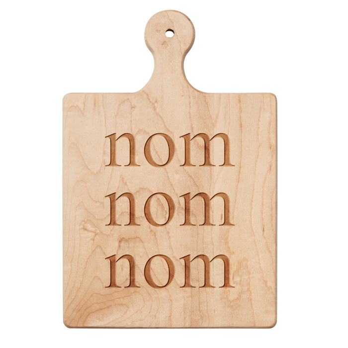 The Maple Artisan Maple Paddle Board | Nom | 9 x 6" features “nom nom nom” carved in three rows—ideal as a playful cheese or charcuterie board at your next gathering.
