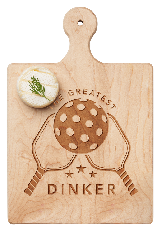 The Maple Artisan Maple Paddle Board | The Greatest Dinker | 9" x 6" features engraved crossed pickleball paddles, a pickleball, and the phrase "The Greatest Dinker." A round cheese with dill sits atop this unique maple cheese board.