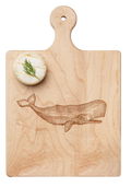 A round cheese with dill rests on the Maple Artisan Maple Paddle Board (Whale, 9" x 6"), engraved with a whale illustration.