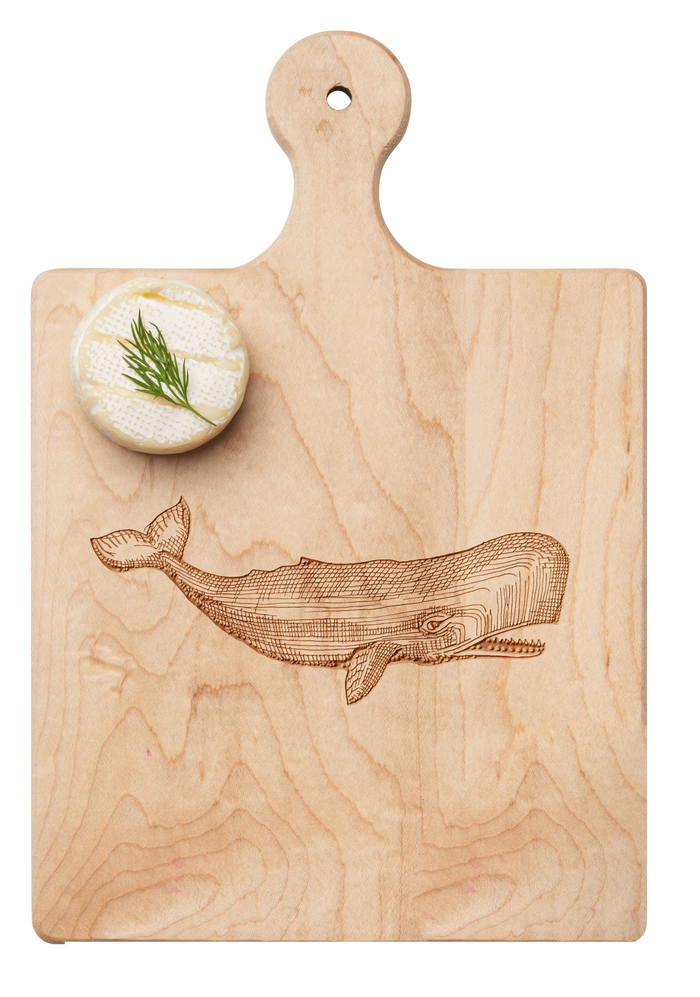 A round cheese with dill rests on the Maple Artisan Maple Paddle Board (Whale, 9" x 6"), engraved with a whale illustration.
