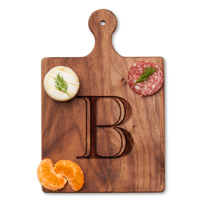 The Maple Artisan Walnut Paddle Board (9 x 6") features a personalized engraved letter B and is styled as a charcuterie board with round cheese, salami topped with dill, and a peeled mandarin orange segment.