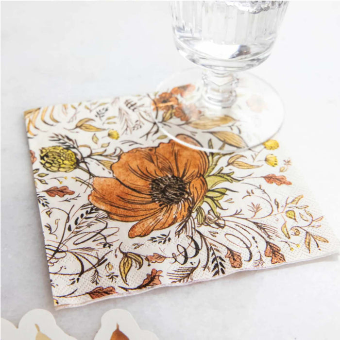 A clear glass rests on Hester & Cook Autumn Arrangement Cocktail Napkins (20pk), featuring an orange floral design with green leaves and yellow accents on a light surface.