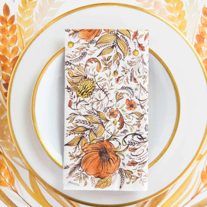 A white plate with a gold rim rests on a matching charger, topped with Hester & Cook Autumn Arrangement Guest Napkins/16pk, featuring orange pumpkins and autumn leaves, displayed on a wheat-themed table.