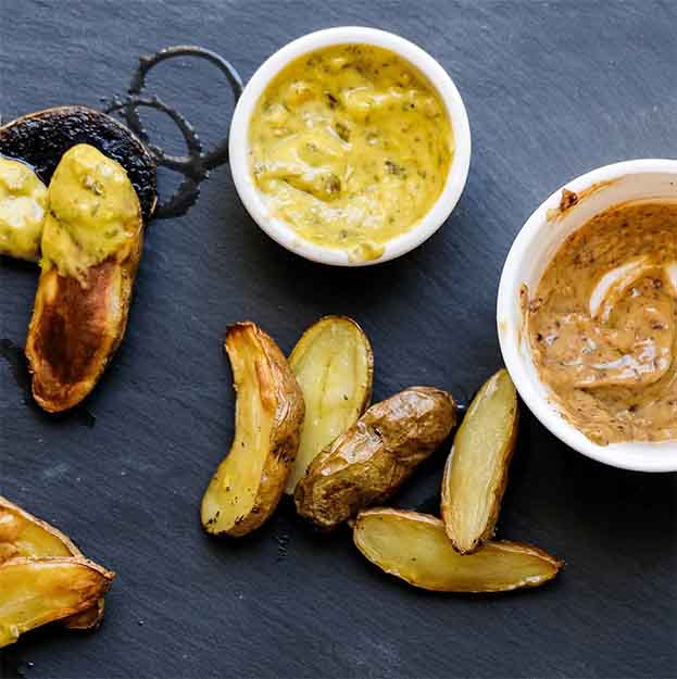 Sliced roasted potatoes on a dark slate surface, served with two dipping sauces—one yellow and creamy, the other Bella Cucina Balsamic Mustard. Perfect for dipping and adding flavor to your next charcuterie board.