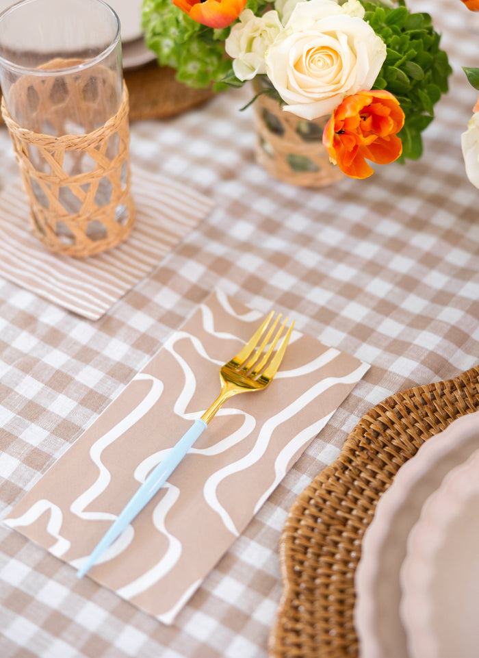 The Sophistiplate Bella Cutlery Gold/Sky Blue Handle/24pkg rests on a patterned napkin atop a beige and white gingham tablecloth, beside a woven placemat, a glass with wicker sleeve, and vivid orange and white flowers.