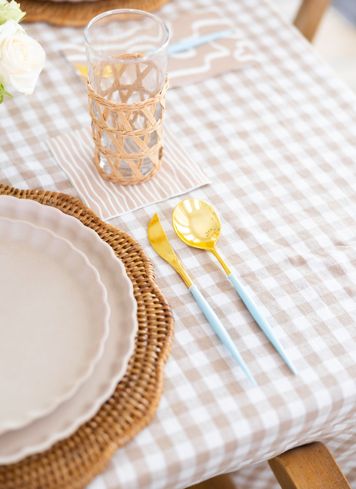 A table setting with a beige scalloped plate, woven placemat, Sophistiplate Bella Cutlery Gold/Sky Blue Handle utensils (24/pkg), a glass in a wicker holder, all on a beige gingham tablecloth.