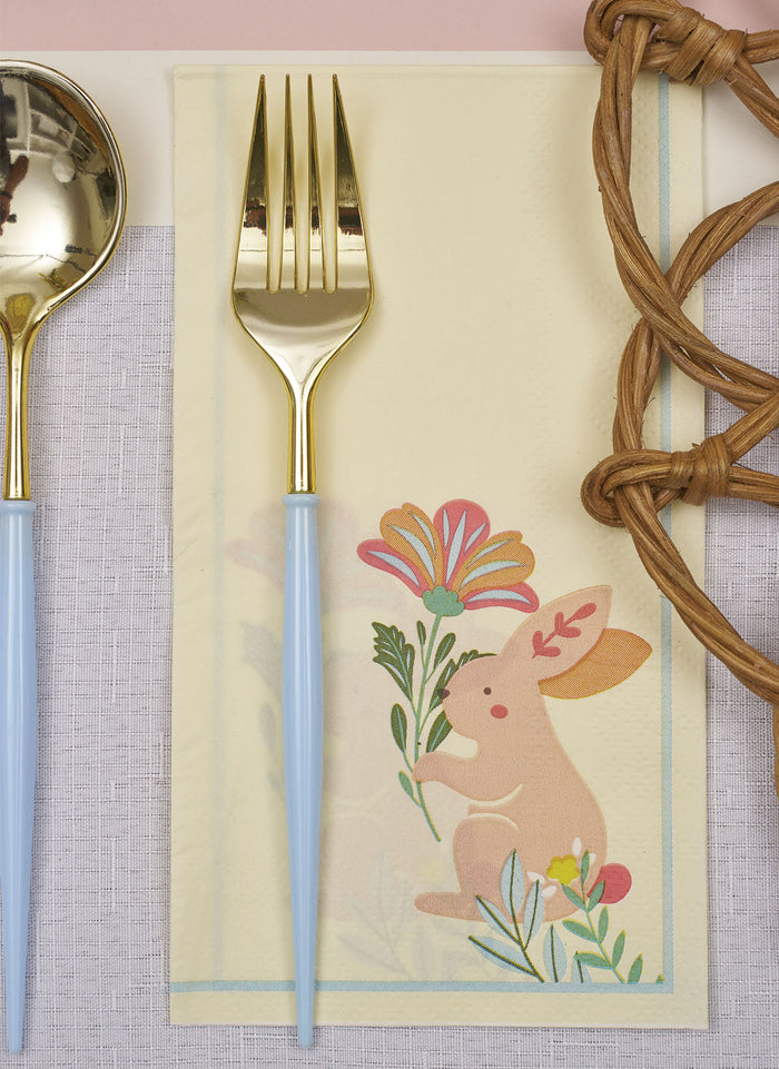 A gold fork with a sky blue handle from the Sophistiplate Bella Cutlery Gold/Sky Blue Handle/24pkg set rests on a bunny-themed napkin, with a matching gold spoon and part of a woven placemat also in view.