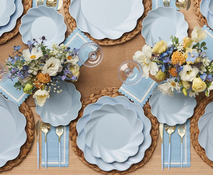 A table set for four features light blue scalloped plates, woven placemats, Sophistiplate Bella Cutlery Gold/Sky Blue Handle, blue napkins, wine glasses, and floral centerpieces with white, yellow, and purple blooms on a burlap runner.