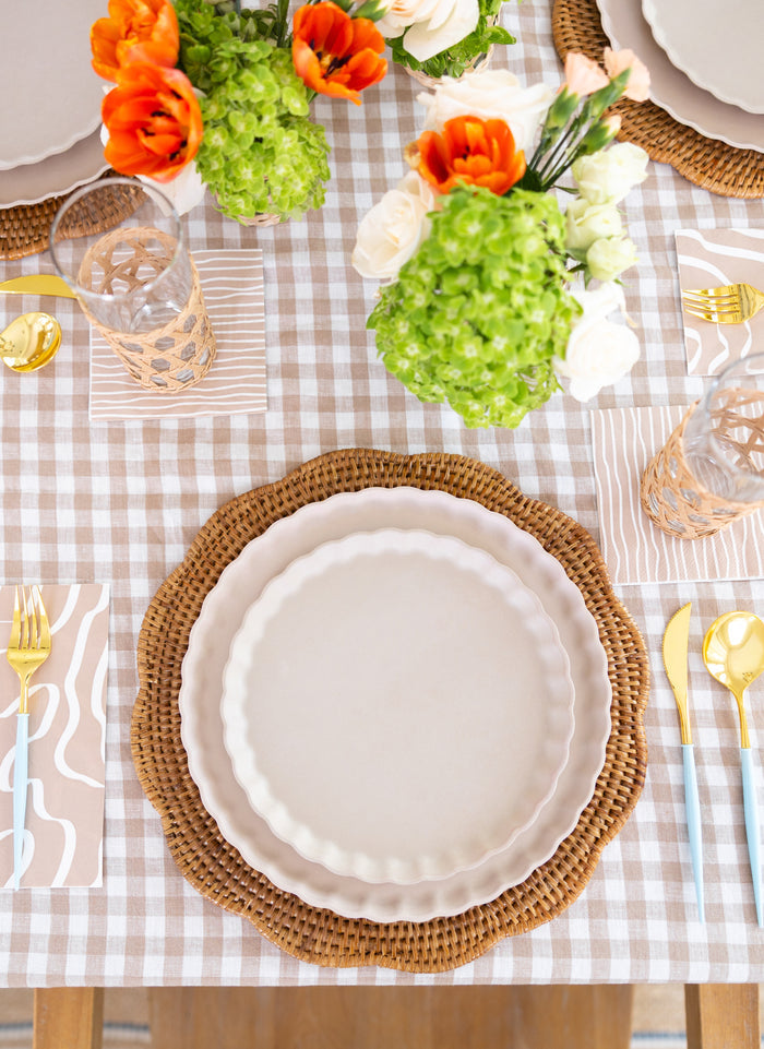 A table is set with white scalloped plates on woven placemats, patterned napkins, glasses, and Sophistiplate’s Bella Cutlery Gold/Sky Blue Handle. A light gingham tablecloth and vases of green and orange flowers complete the look.
