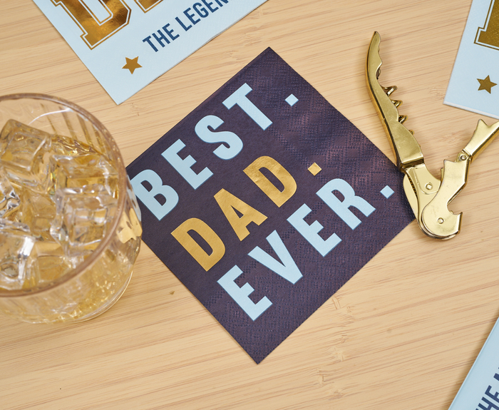 Sugarplum Best Dad Ever Cocktail Napkins (26pk) add a festive touch to Father’s Day, displayed on a wooden surface beside a glass of ice and gold corkscrew—perfect for celebrating dad.