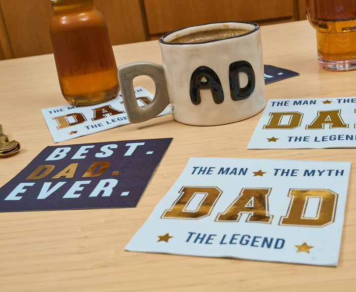 A coffee mug shaped like "DAD" sits on a table with Sugarplum's The Man The Myth Cocktail Napkins (26pk), plus coasters reading BEST. DAD. EVER. and THE MAN THE MYTH DAD THE LEGEND, surrounded by drinks and a bell.