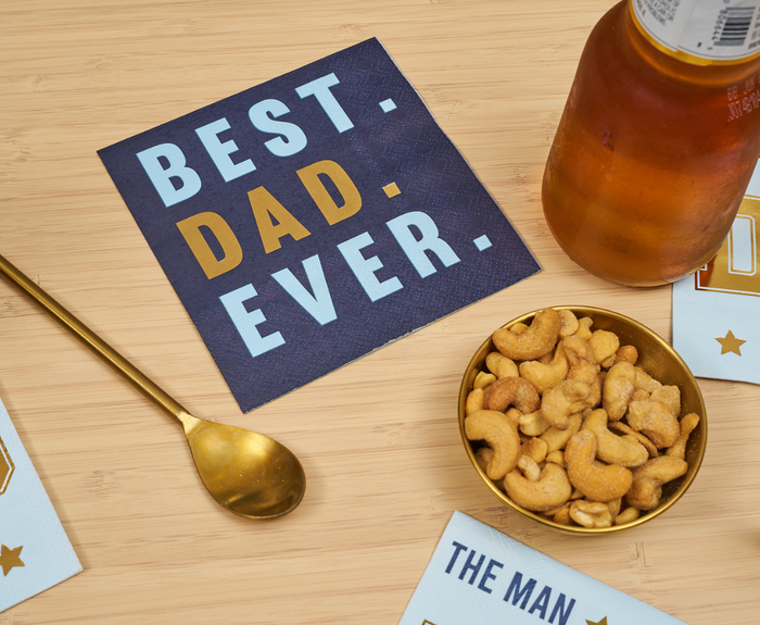 A navy Sugarplum "Best Dad Ever Cocktail Napkins" 26-pack stands out on a wooden table, accompanied by a gold spoon, a bowl of cashews, a beer bottle, and other partially visible napkins.