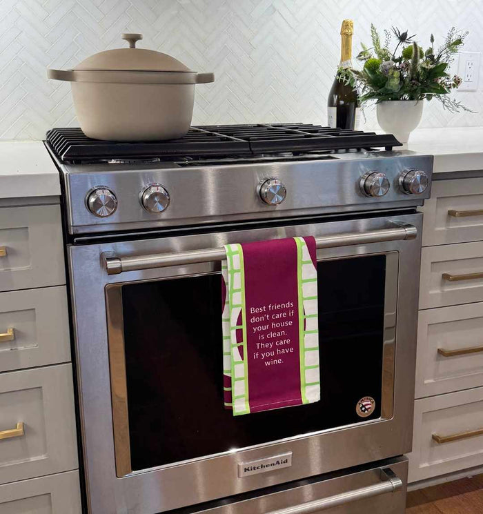 A modern stainless steel oven is accented by the Drinks On Me "Best Friends Dish Towel," which reads, "Best friends don’t care if your house is clean. They care if you have wine," while a pot simmers and wine waits nearby.