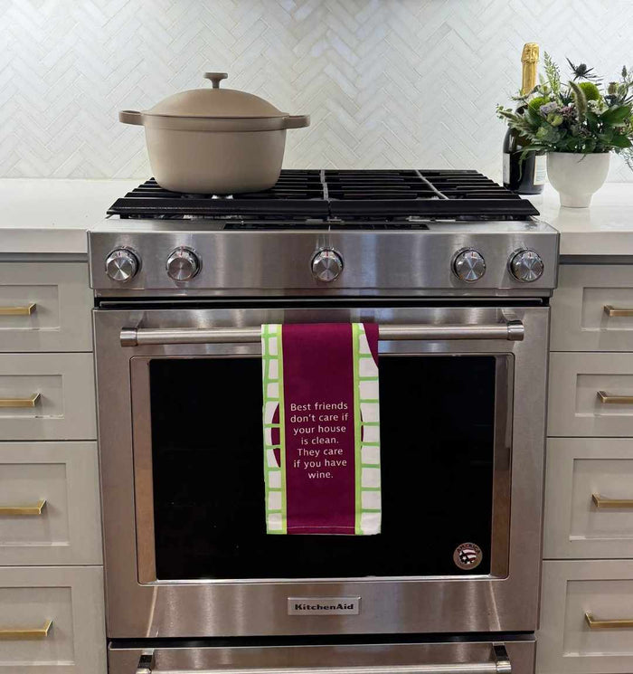 A modern stainless steel oven features the Drinks On Me "Best Friends Dish Towel" draped on the handle, reading, “Best friends don’t care if your house is clean. They care if you have wine.” A beige pot and a flower arrangement are nearby.