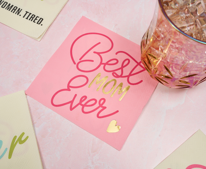 Sugarplum Best Mom Ever Cocktail Napkins (26pk), featuring pink and gold lettering, are perfect for Mother’s Day parties and displayed next to a patterned glass with ice on a light pink surface.