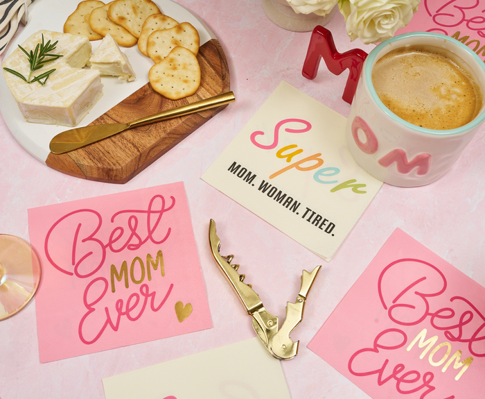 A flat lay featuring Sugarplum Best Mom Ever Cocktail Napkins (26pk), a MOM mug, cheese and crackers, and a corkscrew on a pink surface—perfect supplies for a Mother’s Day celebration.