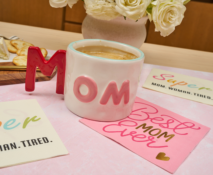 A white mug with pink MOM lettering sits beside cards—Best Mom Ever and Super MOM. WOMAN. TIRED.—with white flowers and Sugarplum Best Mom Ever Cocktail Napkins (26pk) completing the Mother’s Day scene.