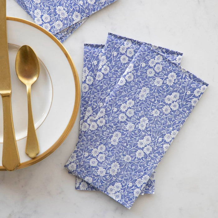A pack of Hester & Cook Blue Calico Paper Guest Towels (16 count) is arranged on a marble surface beside a white plate with a gold rim, topped with an elegant gold fork and spoon.