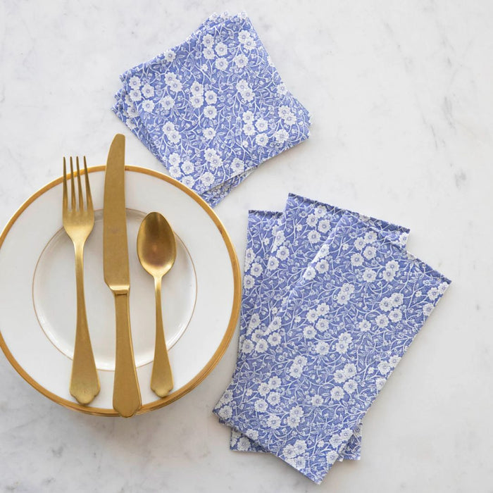 A white plate with a gold rim holds a gold knife, fork, and spoon. Beside it are Hester & Cook Blue Calico Paper Guest Towels (16 pack), some stacked and some spread out on a white marble surface for an elegant table setting.
