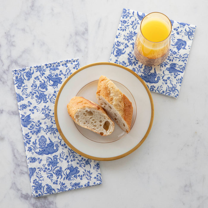 A plate with two pieces of baguette and a glass of orange juice sit on Hester & Cook Blue Regal Peacock Guest Towel Napkins, featuring blue and white floral and peacock designs.