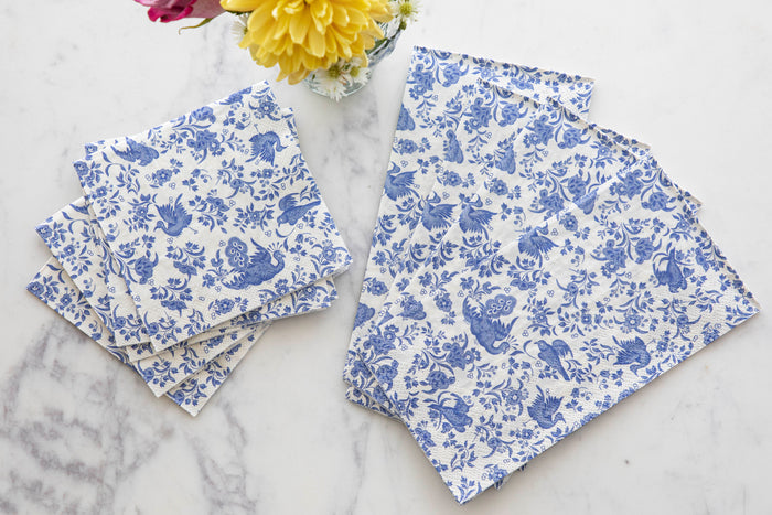 Four folded and three unfolded Blue Regal Peacock Guest Towel Napkins by Hester & Cook, featuring blue floral and animal designs, are arranged on marble. A small vase of yellow and pink flowers adds a charming accent at the top.