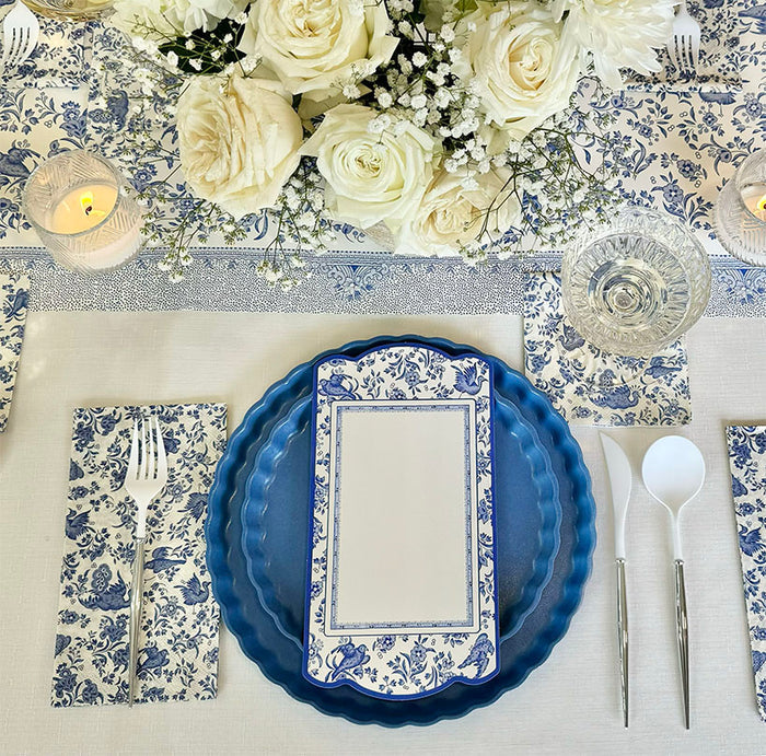 Elegant table setting with blue and white floral-patterned plates, Hester & Cook Blue Regal Peacock Guest Towel Napkins (16pk), a menu card, silverware, glassware, candles, and a bouquet of white roses and baby's breath.